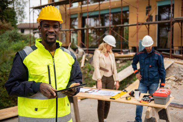 Portrait of man engineer at construction site.Construction manager standing in yellow safety vest and yellow hardhat with crossed arms. Successful confident architect at construction site with team discussing in background. Multiracial Group